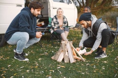 Arkadaşlar piknik için toplandı. Genç adamlar odunları kullanarak şömine yapıyorlar. Kızlar karavanın yanında oturuyor. Çok ırklı insanlar.