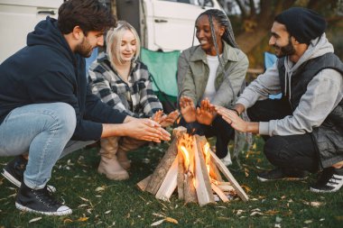 Çok ırklı arkadaşlar şehir dışında piknik yapıyorlar. Ateş yaktılar ve sıcak elleriyle oturdular. Avrupa, siyah, Pakistan ırkı. Minibüsle yolculuk..