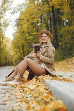 Kadınlar ve köpekler sonbahar parkında oynayıp eğleniyorlar. Kahverengi buldog ve bayan sahibi bir gün parkta oyun oynayıp eğleniyorlar. Bej ceketli ve şapkalı bir kadın..