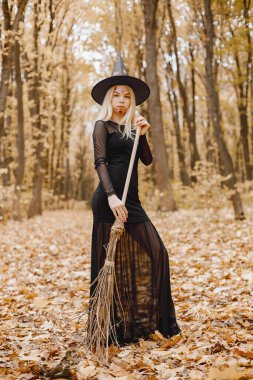 Blonde young girl witch in forest on Halloween. Girl wearing black dress and cone hat. Witch holding a broom.