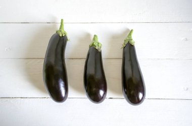 Hafif bir masada üç tane olgun patlıcan var. Beyaz ahşap bir masada üç patlıcan yatıyor..