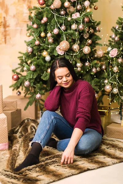 Örgü kazaklı genç bir kadın yılbaşı hediyesiyle bir Noel ağacının yanında yerde oturuyor. Bir kız Noel için bekliyor.