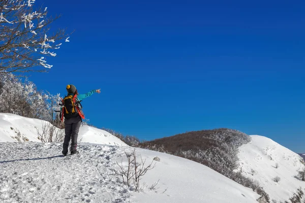Snow hiker in the mountains, with snowshoes and ski poles, backpack and hat. Middle-aged woman points to the surrounding landscape with her hand, full figure.
