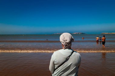 Essaouira, Fas - Agosto 3, 2023: Geleneksel giyimli ve başlıklı bir kadın sahili izliyor. Müslüman dini, kadınların karışık plajlarda çıplak vücutlarını göstermelerini engelliyor..