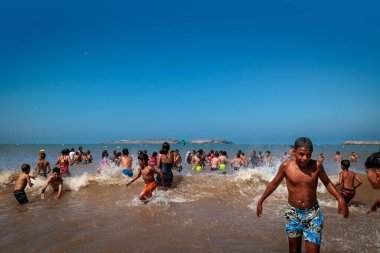 Essaouira, Fas - 3 Ağustos 2023: Bir grup çocuk yüzmek için denize atlıyor, sadece kızlar kendi dinleri tarafından dikte edildiği gibi çok uzun elbiseler veya kostümler giyiyor..