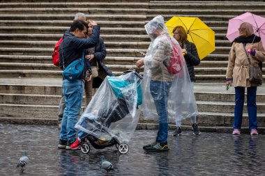 Roma, İtalya - 7 Kasım 2023: Bir sonbahar günü, bir adam oğlunu pusetinde taşır ve şehrin tarihi merkezinde şeffaf bir plastik pelerinle kendini yağmurdan korur..