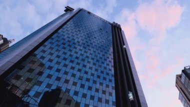 Bulutlu gökyüzünün ve etrafındaki binaların yansıtıldığı bir gökdelenin cephesi. Kapsül şeklindeki dış asansör binanın zeminleri arasında hareket ediyor..