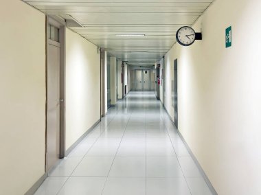 Uzun hastane koridoru, yan kapı ve duvar saati ile. Bir kapı dibinde başka bir alanına giriş işaretleri.