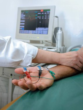 Bir doktor, masada yatan bir hastanın koluna elektrot uygulayarak laboratuvar testleri yapar. Test sonuçlarını izlemek için arka planda elektronik ekipman.