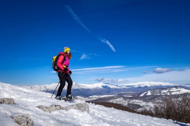 Kar ayakkabılarıyla karda yürüyen aktif sportif bir kadın. Arka planda, kış dağlarının karlı dağları..