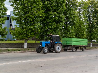Mavi bir traktör yeşil bir römorku sokaktan aşağı çekiyor..
