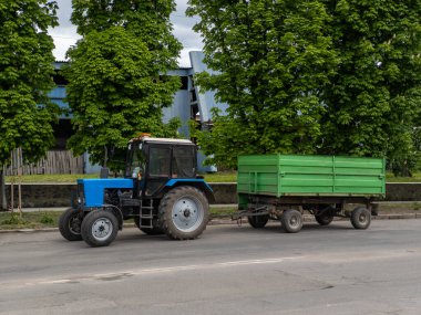 Mavi bir traktör yeşil bir römorku sokaktan aşağı çekiyor..