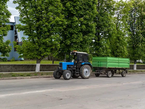 Mavi bir traktör yeşil bir römorku sokaktan aşağı çekiyor..