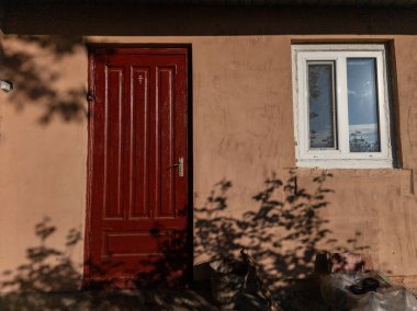 Bir ağacın gölgesi olan pembe bir binanın kırmızı kapısı ve penceresi..