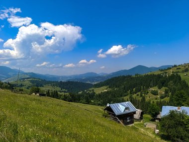 Arka planda dağlar olan çimenli bir tarlanın ortasında küçük bir ev..