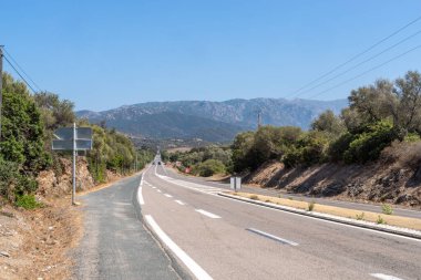 Korsika 'daki dağlık arazide asfalt yol. Sıcak bir gün. Yol boyunca parlak yeşil ağaçlar büyür. Çimler sıcak güney güneşinin altında kurudu. Yüksek kalite fotoğraf