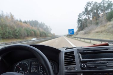 Bir araba, yemyeşil arka planda güzel ağaçlarla, uzun bir yolda sorunsuzca ilerliyor, hoş bir manzara yaratıyor.