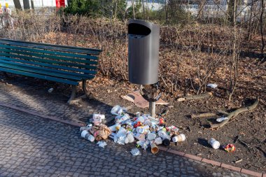 Huzurlu bir park ortamında, çöp tenekesi uygun bir şekilde bir bankın yanına yerleştirilir, atıkların ve çöplerin etkili bir şekilde atılması için bir yer sağlanır.