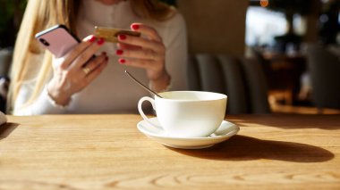 Kafede cep telefonu ve kredi kartı tutan bir kadının elleri. Kahve molası. Masanın üzerinde bir fincan kahve. Bulanık resim, seçici odak