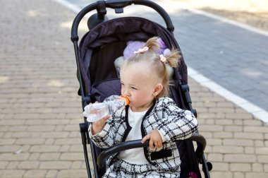 Bebek arabasındaki bebek, yaz parkında yürüyüş yapıyor. Sevimli küçük kız sandalyede oturuyor, elinde bir şişe ile su içiyor. Bebek arabasındaki çocuk