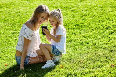 Anne ve kızı yaz parkında çimlerde oturuyor. Akıllı telefon tutan küçük kız, mutlu bir aile uykusu ve iyi vakit geçirin.