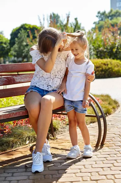 Anne ve kızı güneşli yaz şehir parkında bir bankta oturuyorlar. Çocukluk, eğlence ve insan konsepti. Mutlu aile istirahatı ve iyi vakit geçirmek.