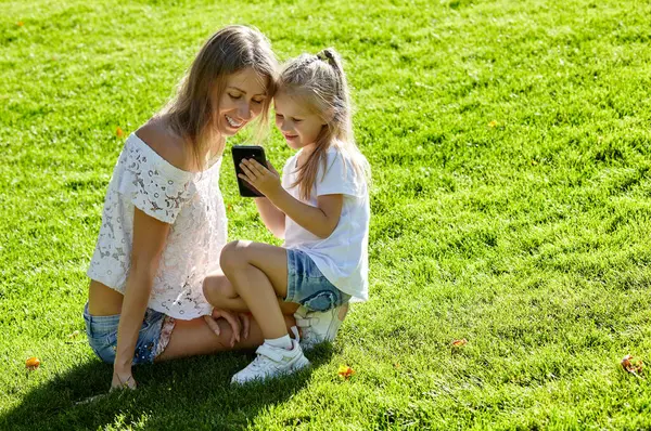 Anne ve kızı yaz parkında çimlerde oturuyor. Akıllı telefon tutan küçük kız, mutlu bir aile uykusu ve iyi vakit geçirin.