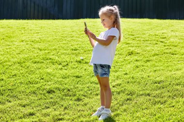 Yaz parkında çimlerin üzerinde duran ve elinde akıllı telefon tutan küçük bir kız. Çocukluk, eğlence ve insan konsepti. Mutlu çocuk istirahati ve iyi vakit geçirin.