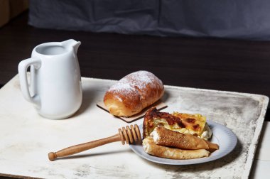 Süzme peynirli ve kuru üzümlü krep ve tabakta süzme peynirli güveç. Reçelli ev yapımı çörek, pudra şekeri ve süt sürahisi mutfak masasında.