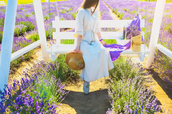 stock image Girl in white linen dress with lavender bouquet and straw hat seat on white bench in Beautiful purple lavender flowers on the summer field. Warm and inspiration concept. Wanderlust and traveling.