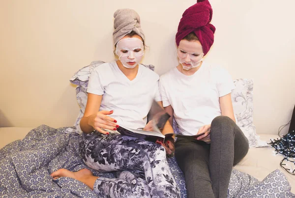 Girls in skin mask sheet on face. Beauty procedures at home. Hen party. Women reading magazine.