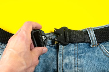 Adamın biri kot pantolonuna siyah bir kumaş kemer iliştirmiş. Koyu renkli metal tokalı modern erkek kemeri..