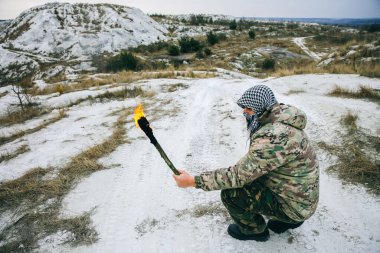 Asker meşaleyi ateşle tutuyor. Kamuflaj üniformalı ve bandanalı bir adam. Dağlarda yaşayan bir adam. Beyaz tepeler ve çöldeki kuru otlar..