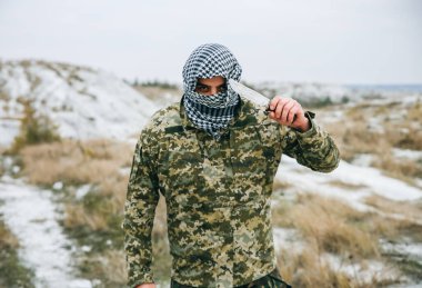 Asker kamuflaj üniforması içinde ve damalı keffiyeh shemagh bandanası içinde duruyor. Bıçaklı adam ıssız bir yerde, dışarıda..