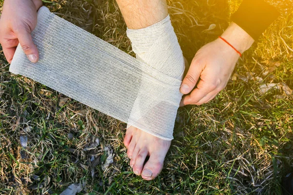 Man is putting bandage on sprained ankle. Twisted leg. Person sitting in the nature. Barefoot on the grass. Dry scratch and wound. Injured tourist. Hiking in uncomfortable shoes. Blister on toe.
