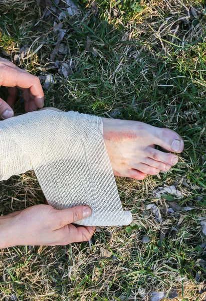 Man is putting bandage on sprained ankle. Twisted leg. Person sitting in the nature. Barefoot on the grass. Dry scratch and wound. Injured tourist. Hiking.