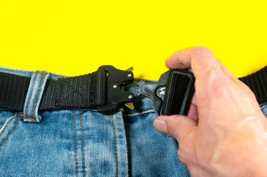 Adamın biri kot pantolonuna siyah bir kumaş kemer iliştirmiş. Koyu renkli metal tokalı modern erkek kemeri..