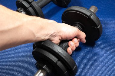 Adam spor salonundaki paspasın üzerinde duran ağır siyah dambılları alır. Fitness sporu motivasyonu. Sağlıklı yaşam tarzınız kutlu olsun. Çubuk ağırlıklarıyla egzersiz. Ev sporu eğitimi.