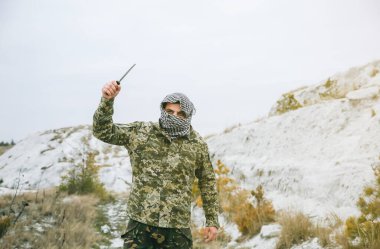 Asker kamuflaj üniforması içinde ve damalı keffiyeh shemagh bandanası içinde duruyor. Bıçaklı adam ıssız bir yerde, dışarıda..
