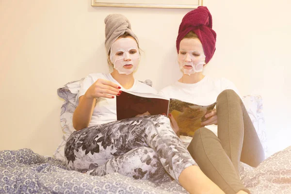 Girls in skin mask sheet on face. Beauty procedures at home. Hen party. Women reading magazine.