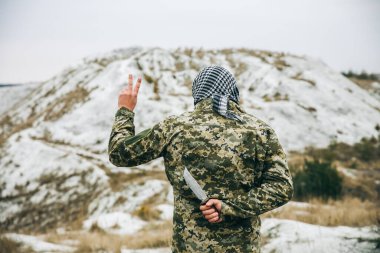 Soldier standing in camouflage uniform and checkered keffiyeh shemagh bandana. Man with knife behind back in the abandoned deserted place. Betraying of enemies. No war and peaceful sign.