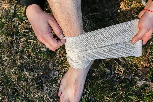 Man is putting bandage on sprained ankle. Twisted leg. Person sitting in the nature. Barefoot on the grass. Dry scratch and wound. Injured tourist. Hiking.