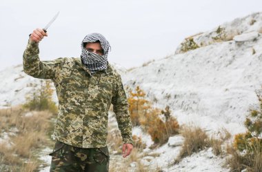 Asker kamuflaj üniforması içinde ve damalı keffiyeh shemagh bandanası içinde duruyor. Bıçaklı adam ıssız bir yerde, dışarıda..