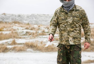 Asker kamuflaj üniforması içinde ve damalı keffiyeh shemagh bandanası içinde duruyor. Bıçaklı adam ıssız bir yerde, dışarıda..