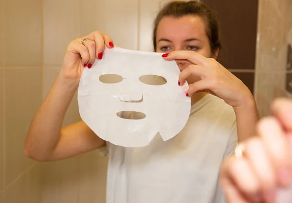 Girl is holding skin mask sheet in hand. Beauty facial procedures at home.