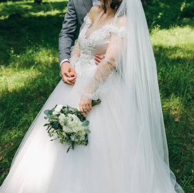 Gelin ve damat parkta sarılıyorlar. Birlikte yürüyen mutlu çift. Düğün günü fotoğrafı. Aşk hikayesi. Güzel uzun kollu elbise. Dantelli peçe. Şık kırsal buket.