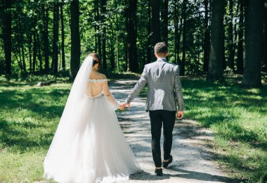 Gelin ve damat parkta el ele tutuşuyorlar. Birlikte yürüyen mutlu çift. Düğün günü fotoğrafı. Aşk hikayesi. Güzel uzun kollu elbise. Dantel peçe.