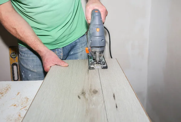 Worker is holding in hands power jigsaw and cutting laminate flooring in apartment. Maintenance repair works renovation with scroll saw. Wooden parquet floor planks indoors.