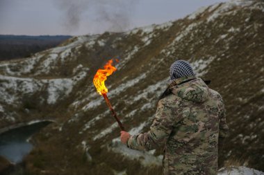 Asker meşaleyi ateşle tutuyor. Kamuflaj üniformalı ve bandanalı bir adam. Dağlarda yaşayan bir adam. Beyaz tepeler ve çöldeki kuru otlar..