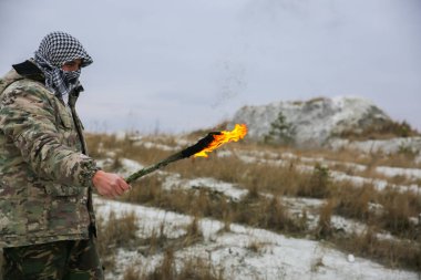 Asker meşaleyi ateşle tutuyor. Kamuflaj üniformalı ve bandanalı bir adam. Dağlarda yaşayan bir adam. Beyaz tepeler ve çöldeki kuru otlar..